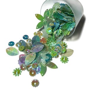 May include: A collection of iridescent green and blue sequins in various shapes, including leaves, flowers, and small circular gears, spilling out from a clear plastic cup. The sequins feature a reflective finish and central holes for attachment.