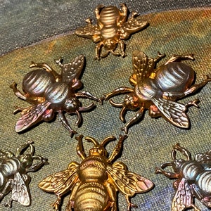 May include: A collection of six metallic bee-shaped decorative items. The bees vary in color from gold to silver, with detailed wings and body segments. The items are arranged on a textured, multi-toned surface.