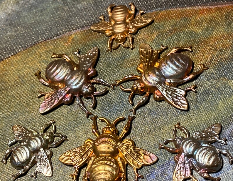 May include: A collection of six metallic bee-shaped decorative items. The bees vary in color from gold to silver, with detailed wings and body segments. The items are arranged on a textured, multi-toned surface.