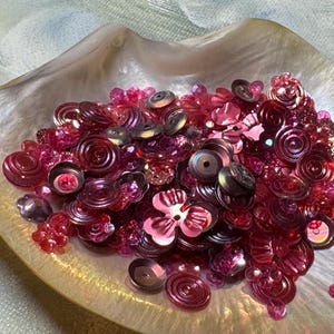 May include: A collection of pink and magenta sequins and beads in various shapes, including flowers and circles, are displayed in a pearlescent, shell-shaped dish. The sequins and beads have a shiny, reflective surface, creating a sparkling effect.