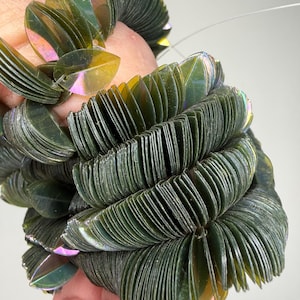 May include: A bracelet composed of multiple dark green, leaf-shaped plastic sequins strung together. Each individual sequin features a subtle iridescent sheen that reflects light, creating a layered, textured appearance along the entire length.