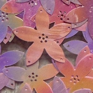 May include: A collection of flat, five-petaled flower shapes in shades of pink, purple, and orange. Each flower features a central cluster of five small circular holes and additional small holes at the tip of each petal, creating a layered pattern.