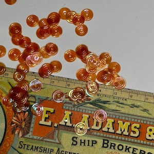 May include: A collection of small, round, orange-tinted translucent sequins with a central hole, scattered across a vintage ruler. The ruler displays metric system markings and the text E.A. ADAMS & CO. SHIP BROKERS, providing a scale reference.