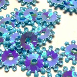 May include: A collection of small, circular, gear-shaped plastic beads featuring a vibrant purple center with a shimmering, iridescent finish and a bright light blue scalloped edge with small, raised circular beads along the perimeter.