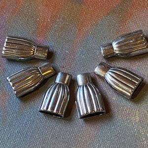 May include: Six silver-colored, ribbed metal clasps arranged in a semi-circle. Each clasp has a cylindrical base and a flared, ridged top. The clasps are likely for jewelry or other craft projects.