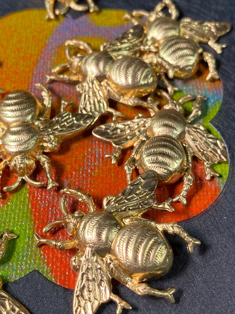 May include: Close-up of several gold-colored bee charms. The bees are detailed with textured wings and bodies. They are arranged on a colorful background of red, green, and blue. The image is well-lit, highlighting the metallic sheen.