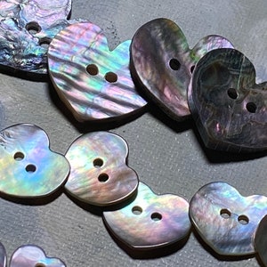 May include: Heart-shaped buttons with iridescent, abalone-like surfaces. Each button has two holes for sewing. The buttons display a range of colors, including blues, greens, and purples, reflecting light. These decorative buttons are ideal for sewing and craft projects.