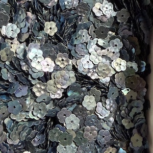 May include: A large collection of small, flower-shaped sequins in a dark, iridescent metallic finish. Each individual sequin features a five-petal design with a single centered hole for attachment, displaying subtle shifts of gray, blue, and silver.
