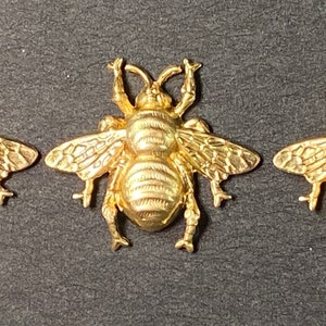 May include: Three gold-colored bee-shaped charms. Each charm has detailed wings, a segmented body, and antennae. The bees are arranged horizontally against a dark background, showcasing their intricate design and metallic sheen.