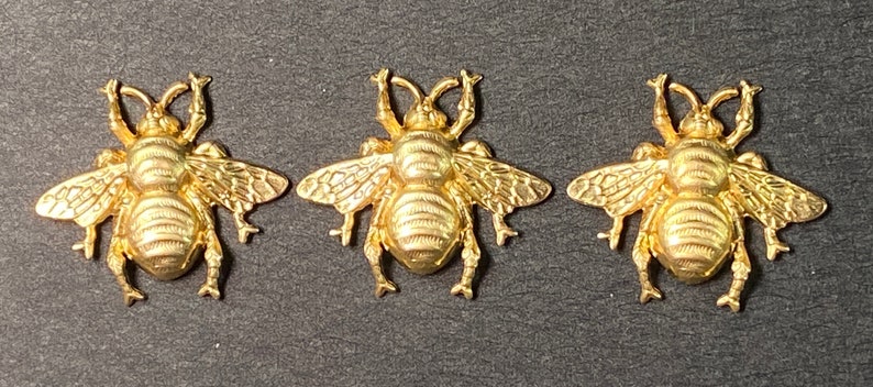 May include: Three gold-colored bee-shaped charms. Each charm has detailed wings, a segmented body, and antennae. The bees are arranged horizontally against a dark background, showcasing their intricate design and metallic sheen.