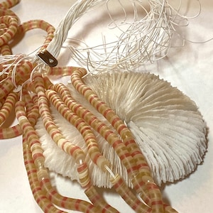 May include: A handmade necklace featuring strands of translucent, striped beads in shades of beige and red. The beads are attached to a white, textured, fan-shaped base. White string and a metal clasp are also visible.