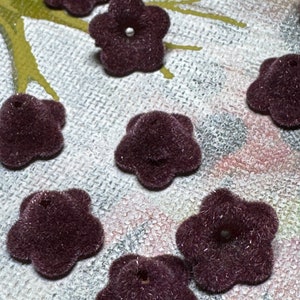 May include: Close-up of several dark purple, flower-shaped embellishments. Each flower has five petals and a small hole in the center. The flowers have a fuzzy texture and a small, silver bead in the center.
