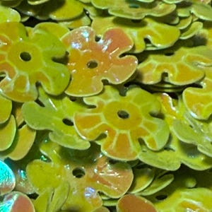 May include: A collection of small, flat, flower-shaped beads with a glossy finish. Each bead features a central hole and a vibrant color palette of lime green and warm orange, with a smooth, reflective surface texture throughout the pile.