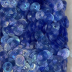 May include: A collection of small, circular, translucent blue sequins. Each individual sequin features a concentric ring pattern with a central hole, creating a layered, textured appearance with varying shades of blue and subtle light reflections.