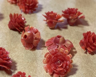 May include: Close-up of several coral-colored flower-shaped beads. The beads have detailed petal designs and a hole through the center for stringing. The beads are scattered on a neutral-toned surface, suggesting a craft or jewelry-making theme.