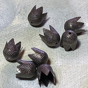 May include: Close-up of several silver-toned metal bead caps, each shaped like a stylized flower with multiple petals. The bead caps have a textured, dotted surface and a small hole at the top for stringing. The caps are scattered on a light-colored surface.