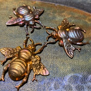 May include: Three metallic bee-shaped brooches in shades of gold and bronze are displayed. The brooches have detailed wings, legs, and antennae. The bees are arranged on a textured, multi-colored surface, creating a striking contrast between the metallic insects and the background.