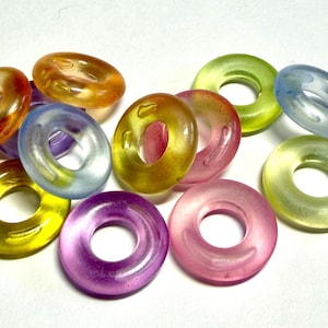 May include: A collection of translucent, ring-shaped beads in various colors, including yellow, orange, pink, purple, green, and blue. The beads are scattered on a white surface, showcasing their glossy texture and vibrant hues. These beads are suitable for jewelry making.