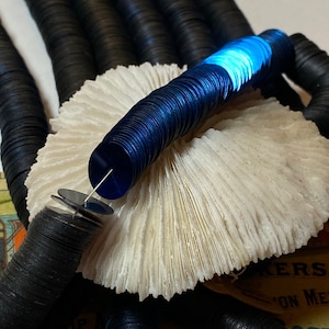 May include: Close-up of black and navy blue sequins, some strung together. The sequins are arranged on a white, textured surface. A few sequins are stacked on a needle. The image has a vintage aesthetic.