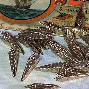 May include: A collection of small, silver-toned metal embellishments, each with a pointed shape and a series of small, decorative rivets. The background includes a vintage nautical-themed illustration with a sailing ship and the text "Steamship Agents".