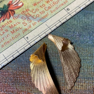 May include: Two gold-colored, wing-shaped earrings with detailed veining. The earrings are set against a vintage background with a fragment of a printed document. The earrings have a textured, metallic finish.