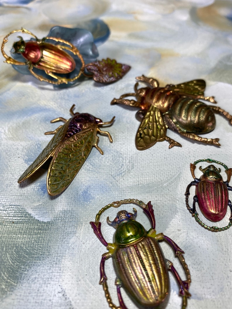 May include: An assortment of decorative insect brooches. The brooches feature detailed designs of beetles, a cicada, and a bee, with metallic gold, green, and red finishes. Some brooches have a ring or halo design around the insect.