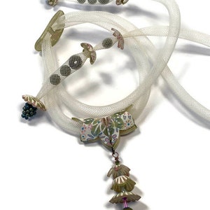 May include: A unique necklace featuring a white mesh cord with decorative elements. The necklace includes floral and geometric accents in shades of green, pink, and brown. A pendant with layered floral shapes hangs from the center, adding a whimsical touch.