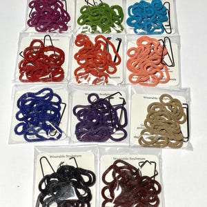 May include: A collection of colorful, coiled rope necklaces in various shades, including red, blue, green, and purple, each packaged with a black paper clip. The packaging reads "Wearable Sculpture".