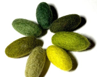 May include: A collection of eight oval-shaped felted wool objects in varying shades of green and yellow, arranged in a circular pattern. The colors range from deep forest green to a bright, almost neon yellow. The objects have a textured, fuzzy appearance.