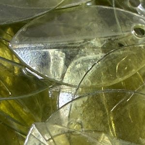 May include: A collection of translucent, pale yellow, leaf-shaped acrylic beads. Each bead features a smooth, curved surface with a distinct central vein and a small, circular hole punched near the top edge for threading, creating a layered effect.