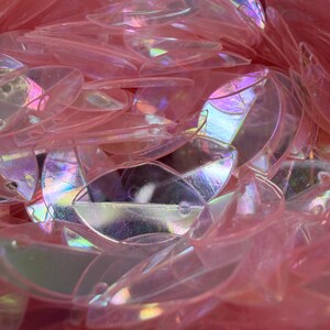 May include: A collection of translucent, light pink, leaf-shaped sequins with an iridescent finish. Each individual sequin features a central vein detail and a small circular hole at one end for attachment, creating a layered, reflective surface.