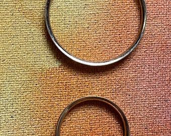 May include: Two silver-colored metal bangles, one larger than the other, are displayed against a textured, golden-brown background. The bangles are circular and appear to be made of a shiny metal.