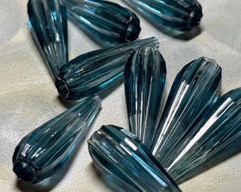 May include: A collection of teal-colored, teardrop-shaped glass beads. Each bead has a ribbed texture and a hole at the top for stringing. The beads are scattered on a light-colored surface, creating a play of light and shadow.
