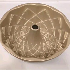 Puede incluir: Un molde para bizcocho bundt dorado con un diseño geométrico. El molde tiene un tubo central y un patrón decorativo de formas de diamante y triangulares. El molde es de metal y está diseñado para hornear pasteles.