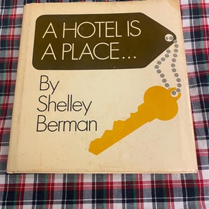 Può includere: Un libro vintage intitolato "A Hotel Is A Place..." di Shelley Berman. La copertina è bianco sporco con una grafica a etichetta marrone e un'illustrazione di chiave gialla. Lo sfondo è un motivo a quadri rosso, bianco e blu.