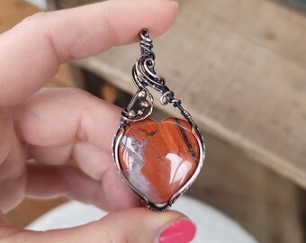 May include: A heart-shaped red and white stone pendant with copper wire wrapping. The pendant is held by a hand.