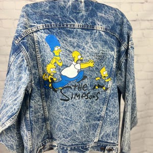 Puede incluir: Chaqueta vaquera con lavado ácido con un gráfico colorido de la familia Simpson en la espalda. La chaqueta tiene un cuello clásico, cierres de botón y un aspecto vintage. El logotipo de los Simpsons está impreso debajo.