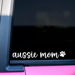 May include: White vinyl decal with the words "aussie mom" in a cursive font, followed by a paw print. The decal is displayed on a vehicle window, with a pink door frame visible.