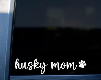 May include: A white vinyl decal with the text "husky mom" and a paw print on a black background. The decal is applied to the back window of a pink car.