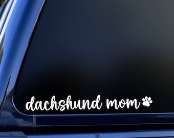 May include: White vinyl decal with the text "dachshund mom" and a white paw print on a black car window.
