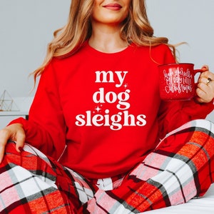 May include: A red sweatshirt with the text "my dog sleighs" in white. The person wearing the sweatshirt is also wearing red and black plaid pajama pants.