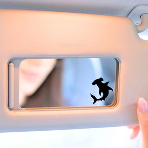 May include: A black hammerhead shark decal is affixed to a car visor. The visor is a light beige color, and the background shows a blurred reflection. The shark is a silhouette, with its distinctive head shape clearly visible.