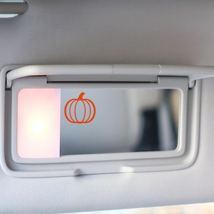 May include: A car visor with a built-in mirror and a small orange pumpkin decal. The visor is gray and has a light on the left side. The pumpkin is a simple outline design.