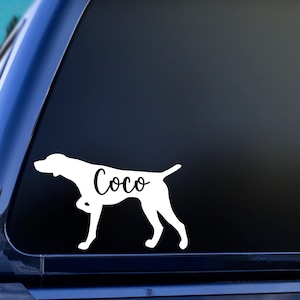 May include: White vinyl decal of a German Shorthaired Pointer dog silhouette with the name "Coco" written on the dog's side. The decal is on a blue car window.