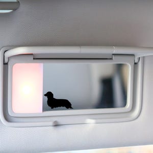 May include: A black silhouette of a long-haired dachshund dog is affixed to a car visor mirror. The visor is white and has a built-in light. The background is out of focus.