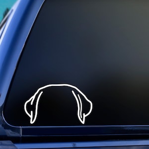 May include: White vinyl decal of a dog's head, showing the ears and top of the head. The decal is a simple line drawing, suitable for car windows or other smooth surfaces. The design is a minimalist representation of a canine.