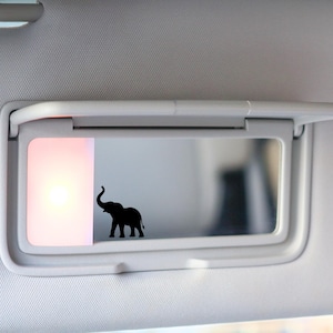 May include: A black elephant silhouette decal is affixed to the mirror of a car visor. The visor is light gray, and the mirror is reflecting a blurred interior. The light is on, illuminating the mirror.