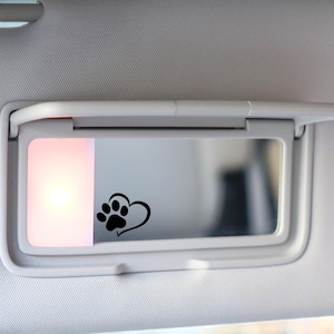 May include: A car visor with a built-in mirror. A black paw print and heart design is on the mirror. The visor is white and has a light on the left side.