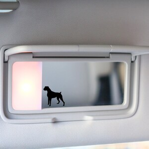 May include: A car visor with a built-in mirror and a black silhouette of a dog. The visor is white and has a rectangular light on the left side. The dog silhouette is in the mirror.