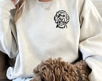 May include: A light beige sweatshirt featuring a black graphic of a dog's face on the upper left chest. The dog has curly fur and a friendly expression. The sweatshirt is a neutral color and has long sleeves.